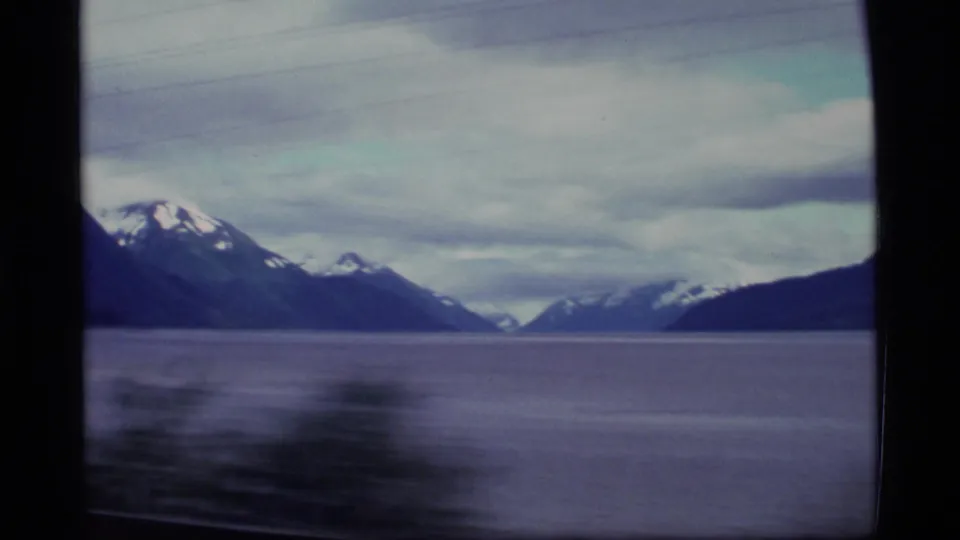1977 Alaska 8mm travel view of calm water and distant snow-capped mountains framed by a dark window edge.