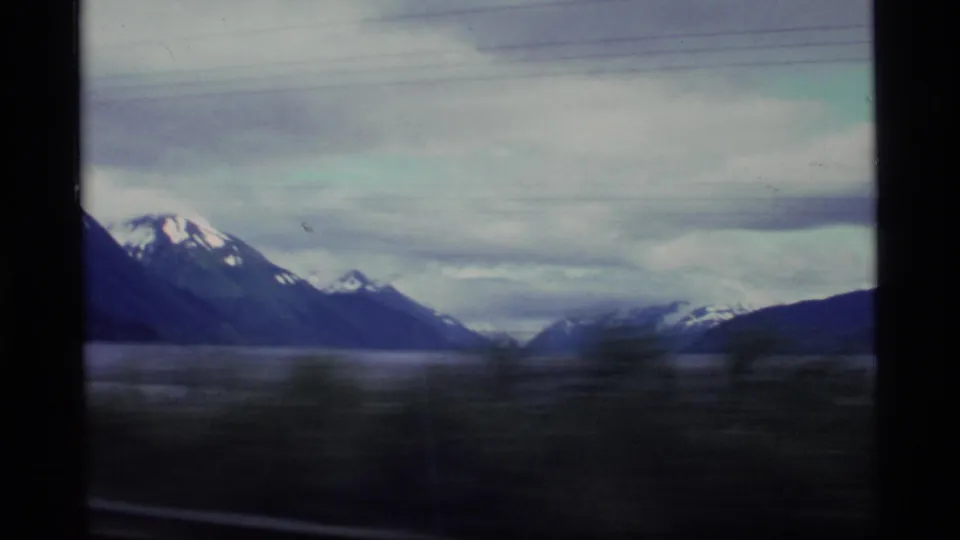 1977 Alaska home-movie frame showing water, snow-topped mountains, and blurred foreground movement from a vehicle.