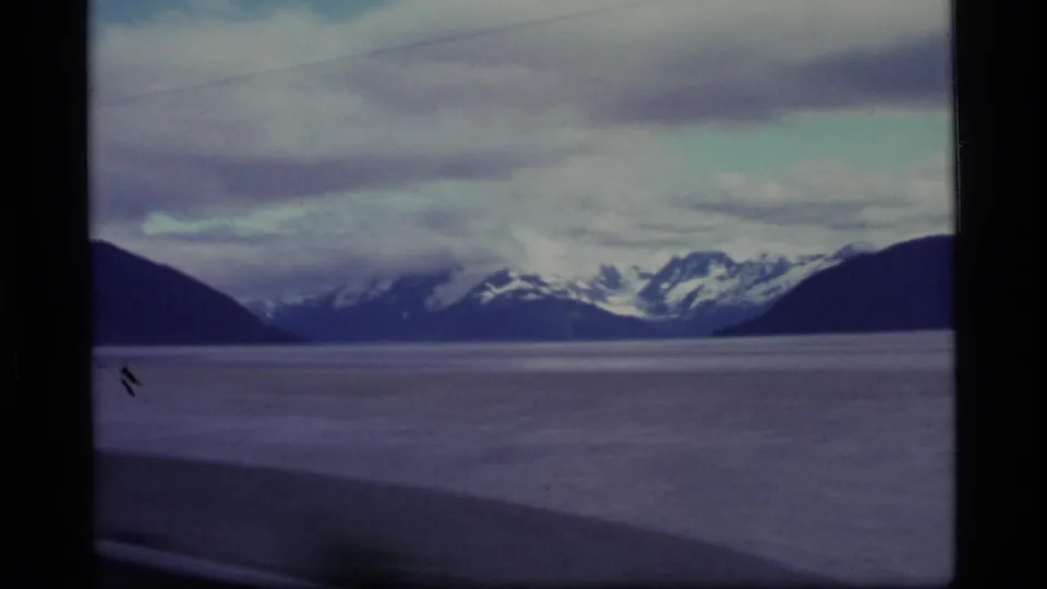 1977 Alaska 8mm scenic frame of a wide body of water bordered by dark slopes and snowy mountains.