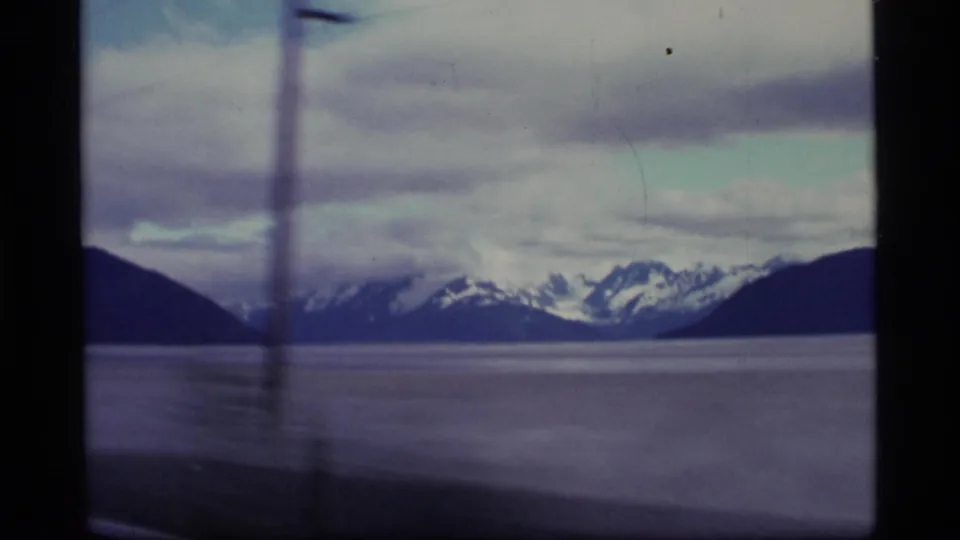 1977 Alaska travel frame with distant snowy mountains, calm water, and a blurred roadside utility pole in front.