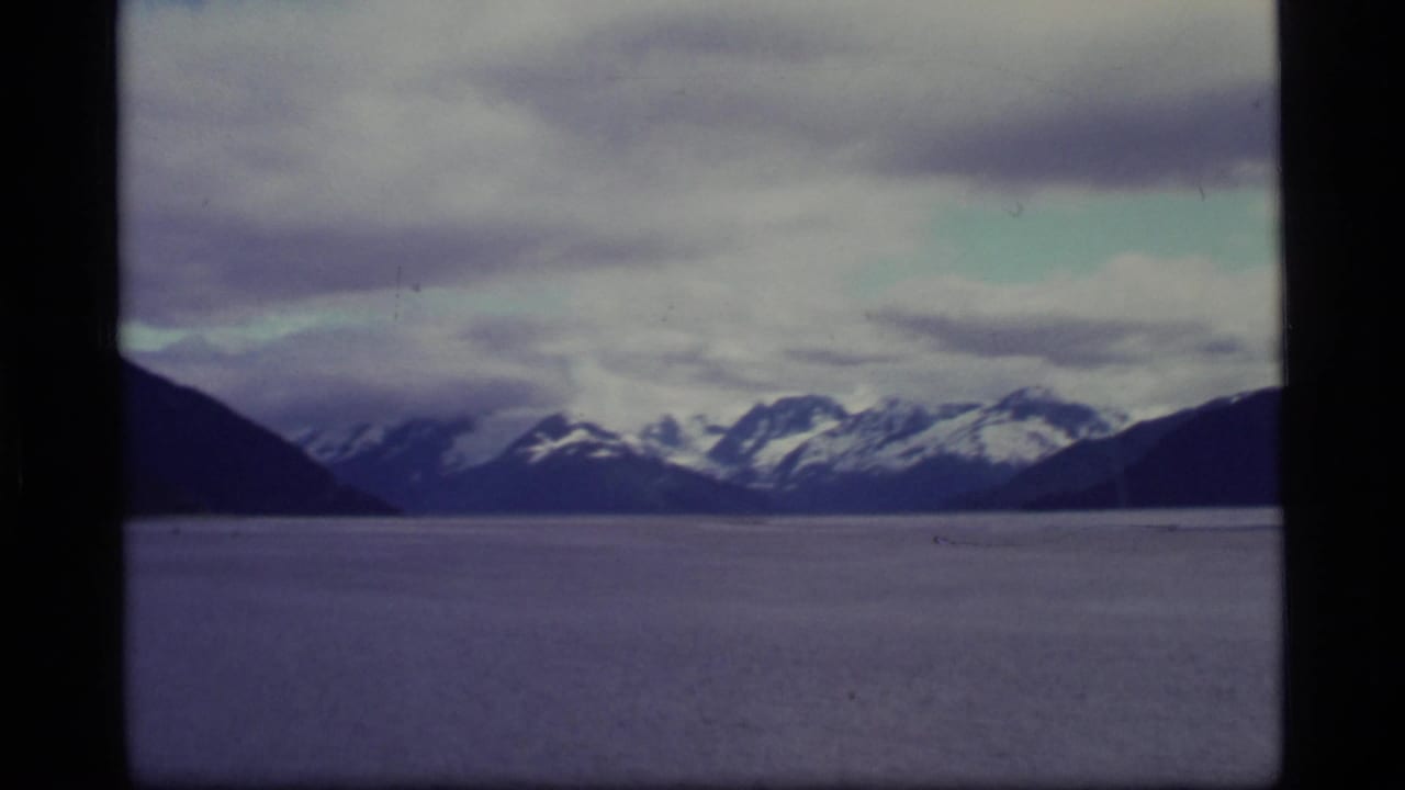 1977 Alaska 8mm frame showing distant snow-capped mountains across a broad body of water beneath layered clouds.