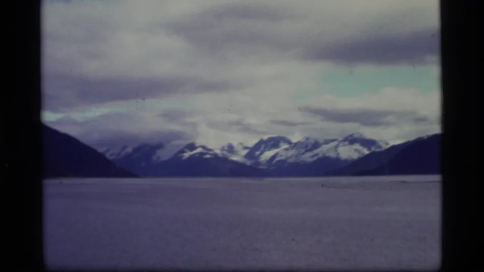 1977 Alaska 8mm frame showing distant snow-capped mountains across a broad body of water beneath layered clouds.