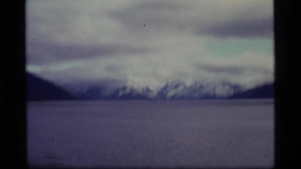 Soft 1977 Alaska home-movie view of snowy mountain ridges beyond water, partially bordered by dark film or window edges.