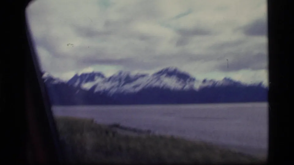 1977 Alaska 8mm frame with snow-streaked mountains across water and a grassy shoreline foreground, likely filmed from near or inside a vehicle.