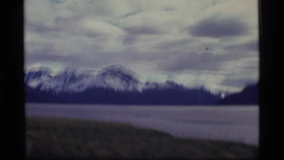 1977 Alaska home-movie frame showing a cloudy mountain landscape across water with a low shoreline foreground and visible 8mm softness.