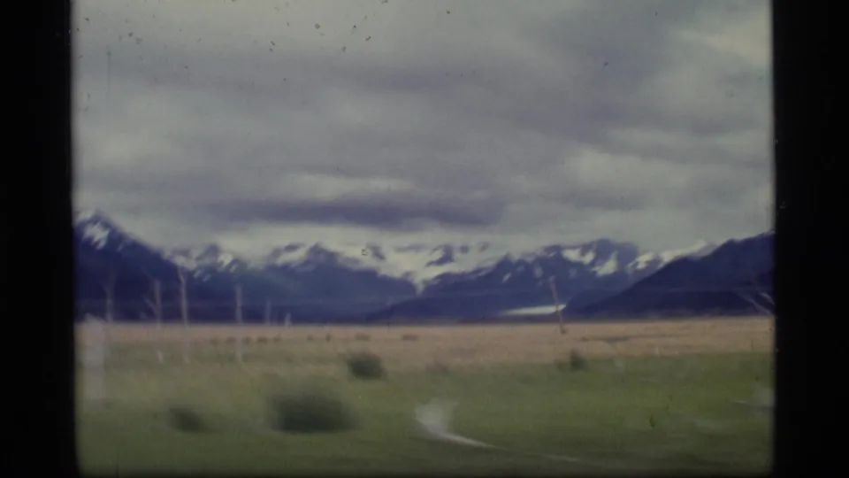 1977 Alaska 8mm view of distant snowy mountains and open grassland beneath layered clouds, with visible film gate borders.