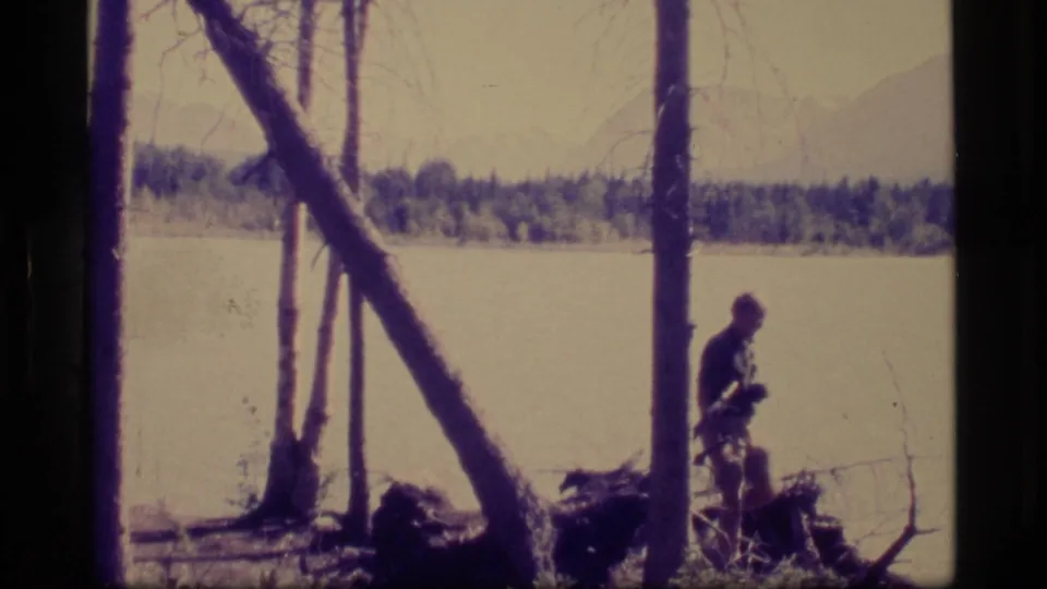 1977 Alaska lakeside home-movie frame with a distant person walking beside bare trees, water, forest, and mountains beyond.