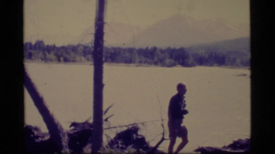 1977 Alaska 8mm frame showing a lone lakeshore walker, foreground trees, pale water, distant forest, and hazy mountains.