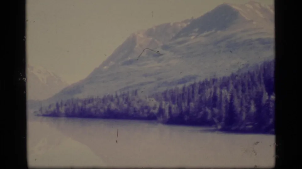 1977 Alaska 8mm frame showing a large mountain, tree-covered shoreline, and calm water with a soft reflected shape near the bottom edge.
