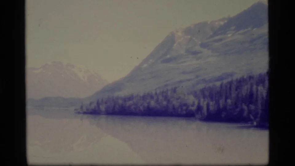 1977 Alaska 8mm frame of a mountain above a wooded shoreline and calm lake or river water, with faded color and soft focus.