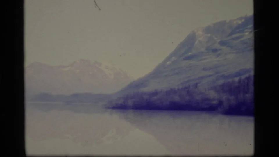 1977 Alaska 8mm frame showing snow-capped mountains, dark shoreline forest, and a calm lake with soft reflections.