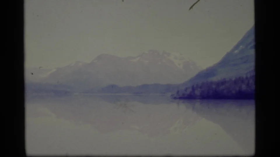 1977 Alaska 8mm frame with a distant snow-covered mountain range, quiet lake surface, and forested shore at the right edge.