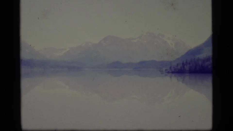 1977 Alaska 8mm frame showing a broad lake, hazy mountain backdrop, forested shoreline, and subdued reflections in the water.