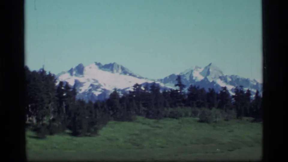 1977 Alaska 8mm frame with distant snow-capped mountains rising over evergreen forest and open green meadow.