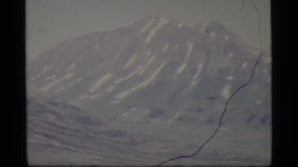 1977 Alaska 8mm frame showing a distant snow-streaked mountain ridge under a pale sky with faded color and film wear.