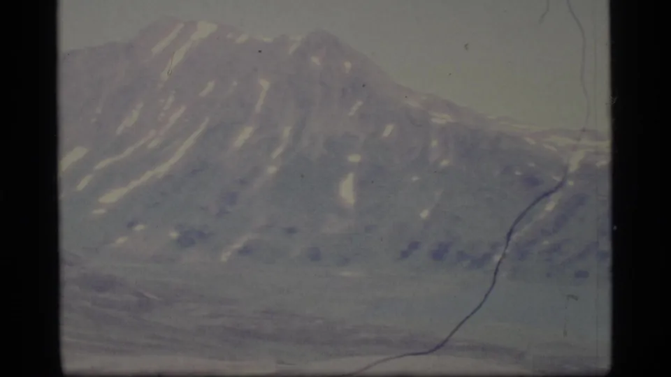 1977 Alaska 8mm frame of a rugged mountain face with scattered snow patches, soft focus, faded color, and visible scratches.