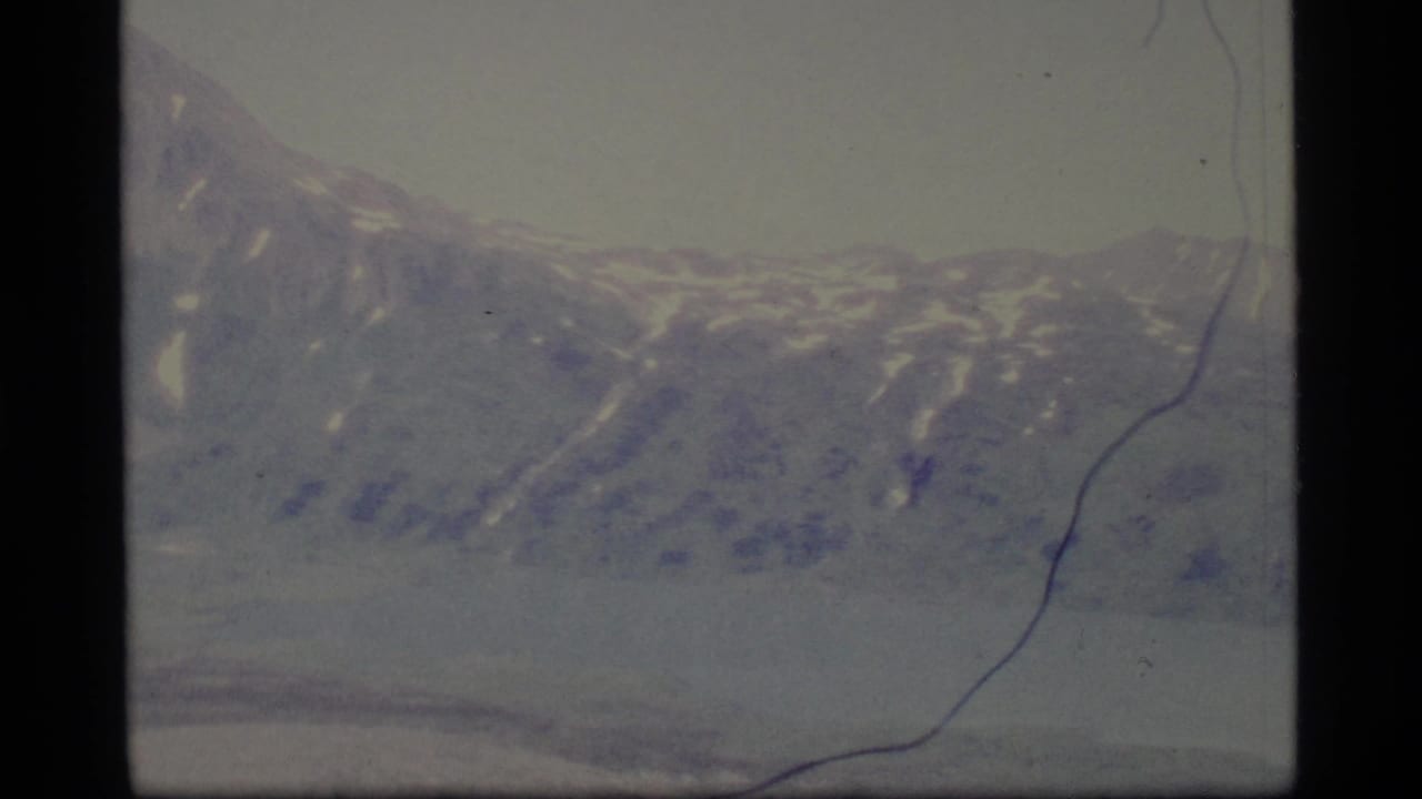 1977 Alaska 8mm frame showing distant snow-dusted mountains across a calm lake under a hazy sky.