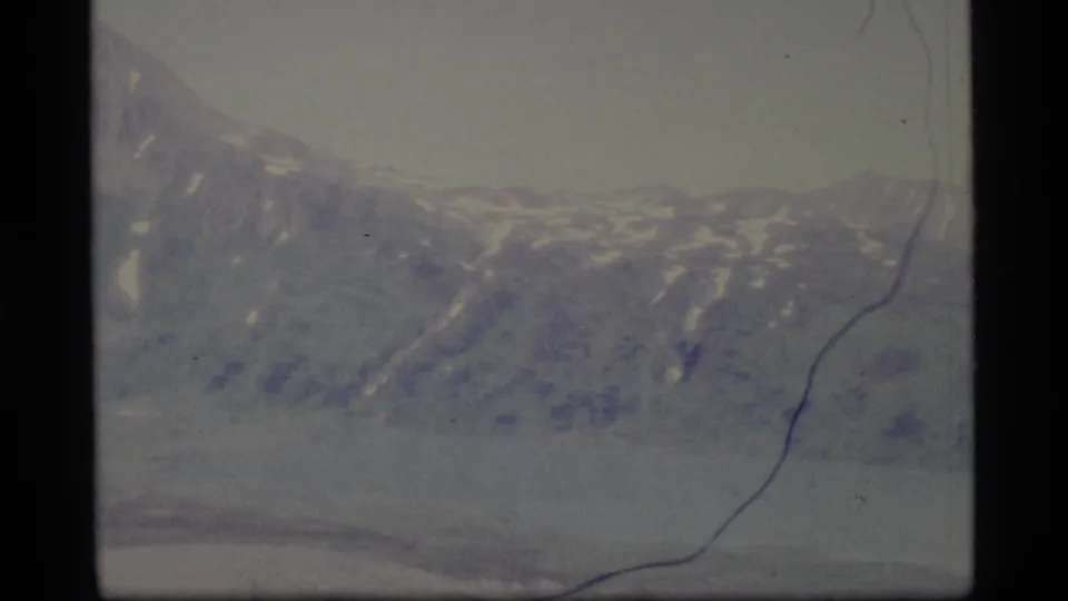 1977 Alaska 8mm frame showing distant snow-dusted mountains across a calm lake under a hazy sky.