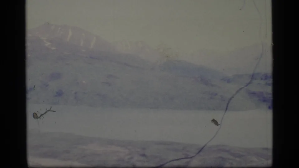 1977 Alaska 8mm view of a hazy mountain ridge beyond pale lake water with visible frame scratches.