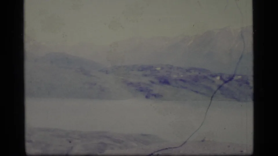 1977 Alaska 8mm frame showing a broad mountain range above a lake, with muted color and visible film wear.