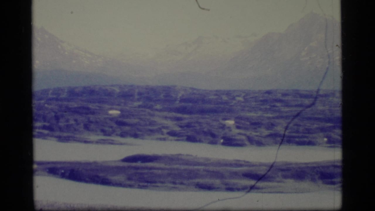 Snowy mountain range beyond blue-purple icy terrain and water, with film scratches and dark frame borders.