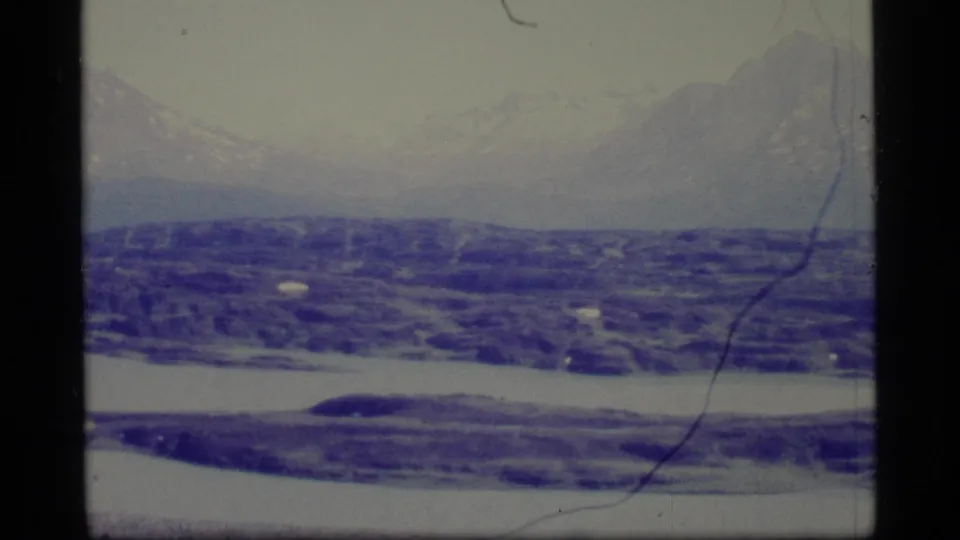 1977 Alaska 8mm frame showing distant snow-capped mountains above blue-purple ice or water, with visible grain, scratches, and soft exposure.