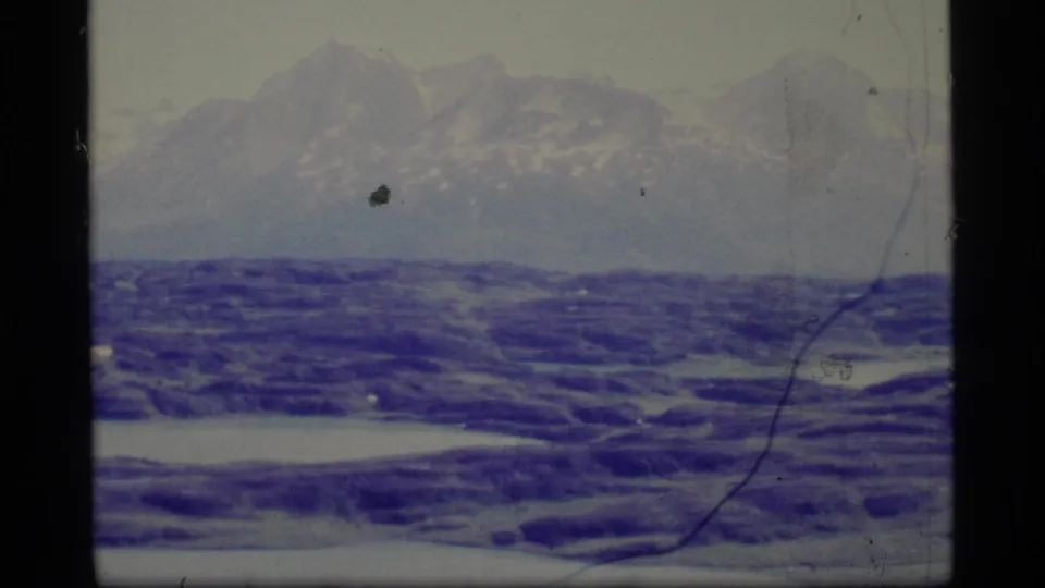 1977 Alaska 8mm frame with a broad mountain backdrop, blue-purple ice or water in the foreground, and visible dust and scratch marks.
