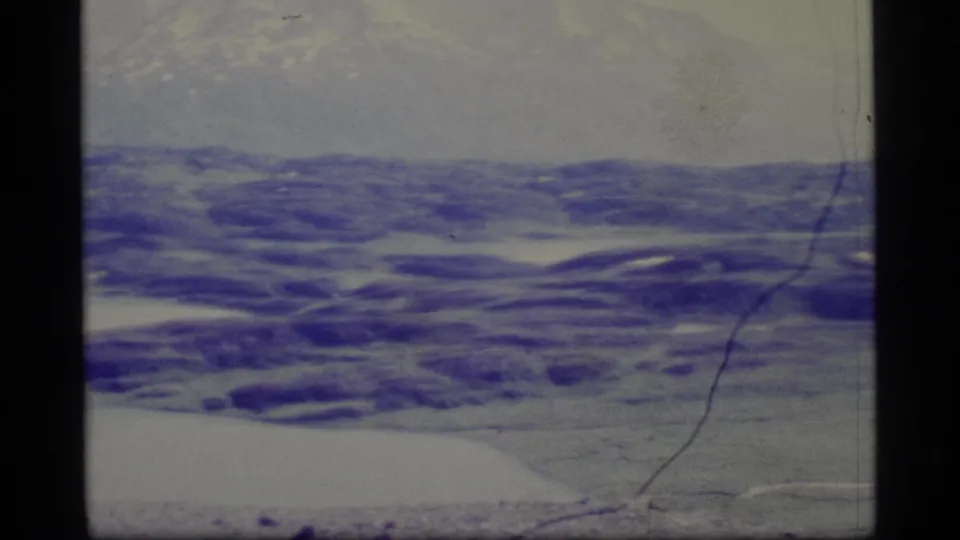 1977 Alaska 8mm frame showing a low, remote icy landscape with distant mountains, pale sky, and visible scratches from the physical film scan.