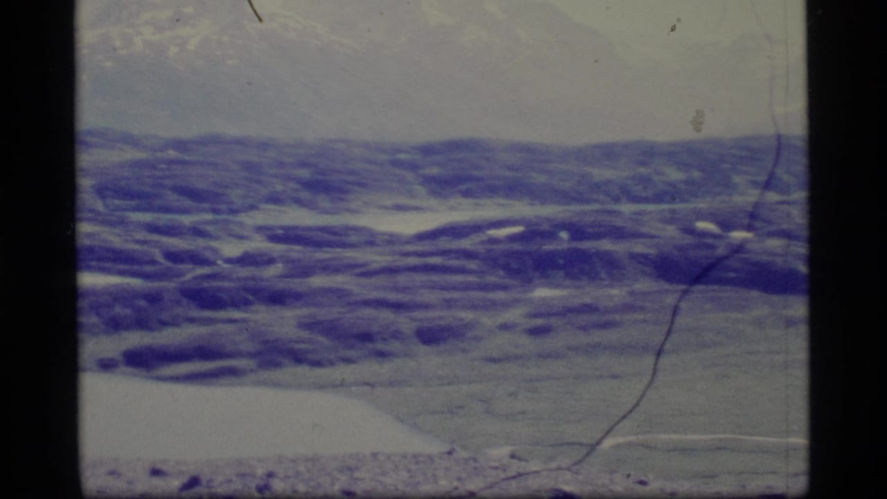 1977 Alaska 8mm aerial frame showing snow-streaked hills and distant terrain with aged blue-purple color shift.