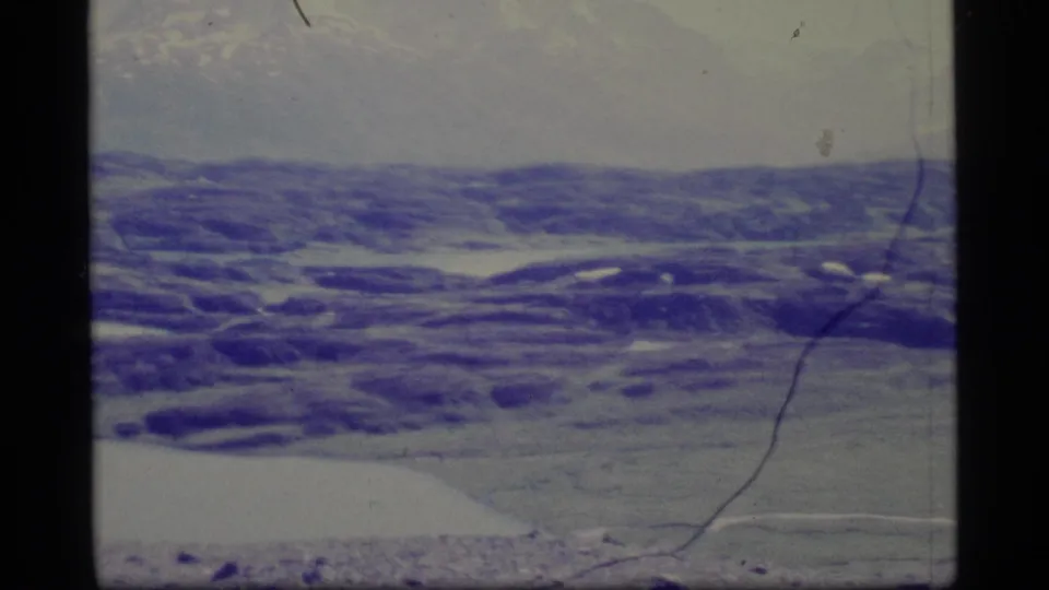 1977 Alaska 8mm aerial frame showing snow-streaked hills and distant terrain with aged blue-purple color shift.