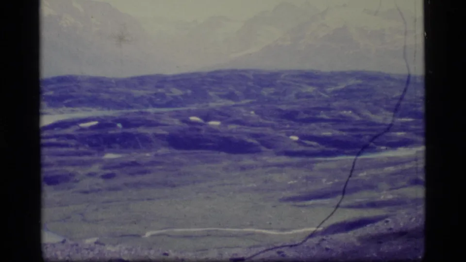 1977 Alaska home-movie frame showing a soft aerial view of low snowy hills and open terrain through faded film color.