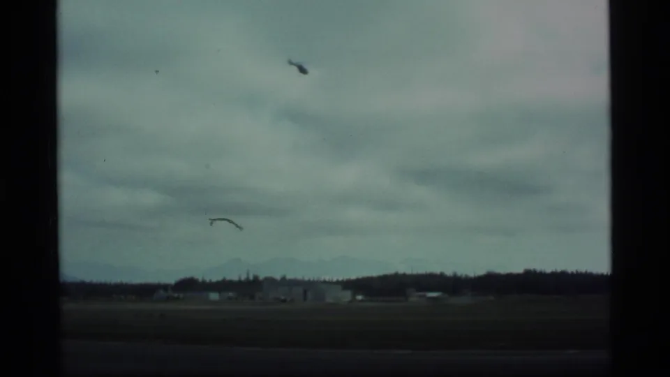 Alaska 1977 8mm frame showing open ground, a wooded horizon, distant low structures, and a cloudy sky.
