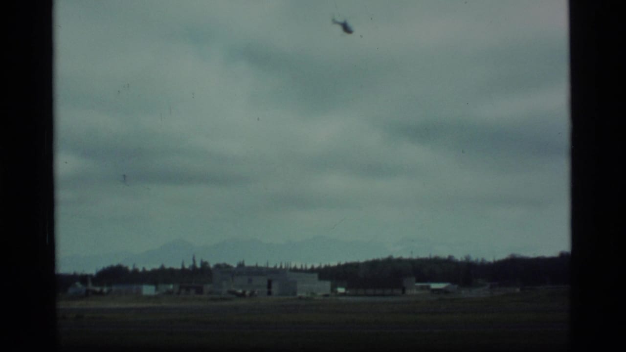 1977 Alaska home-movie frame showing low airfield buildings, a dark runway area, cloud cover, and faint mountains in the distance.