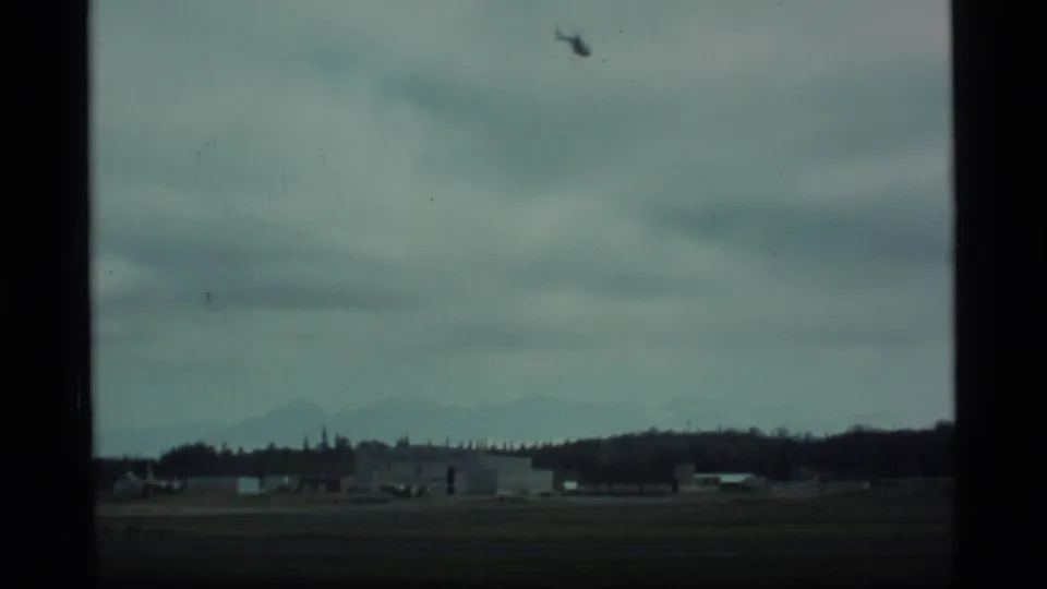 1977 Alaska home-movie frame showing low airfield buildings, a dark runway area, cloud cover, and faint mountains in the distance.
