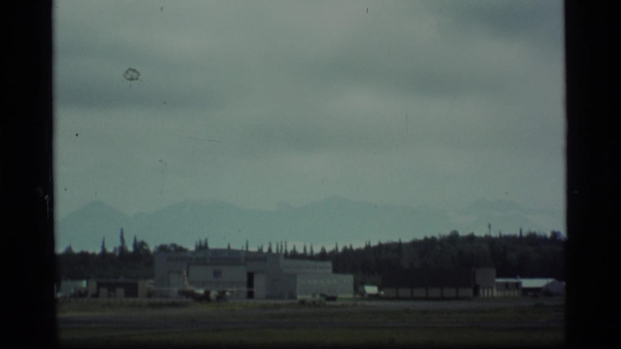Airfield hangar and nearby structures with mountains beyond the runway area.