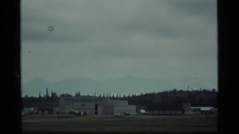 1977 Alaska airfield view with a hangar-like building, low structures, tree line, and hazy mountains beneath overcast sky.