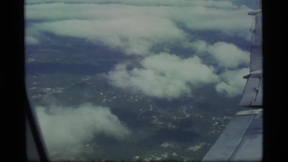 1977 Alaska aerial home-movie view looking past an aircraft wing over scattered clouds, roads, and dark terrain below.