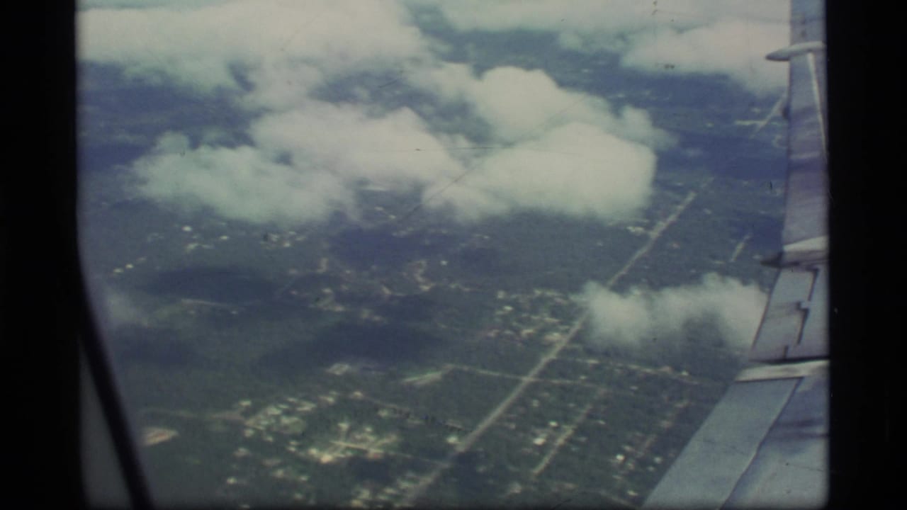 Aerial view through an airplane window with a wing edge, clouds, roads, and green terrain below.