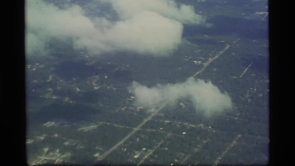 1977 8mm Alaska flight view showing clouds over green land, faint road lines, and scattered settlement or cleared areas.