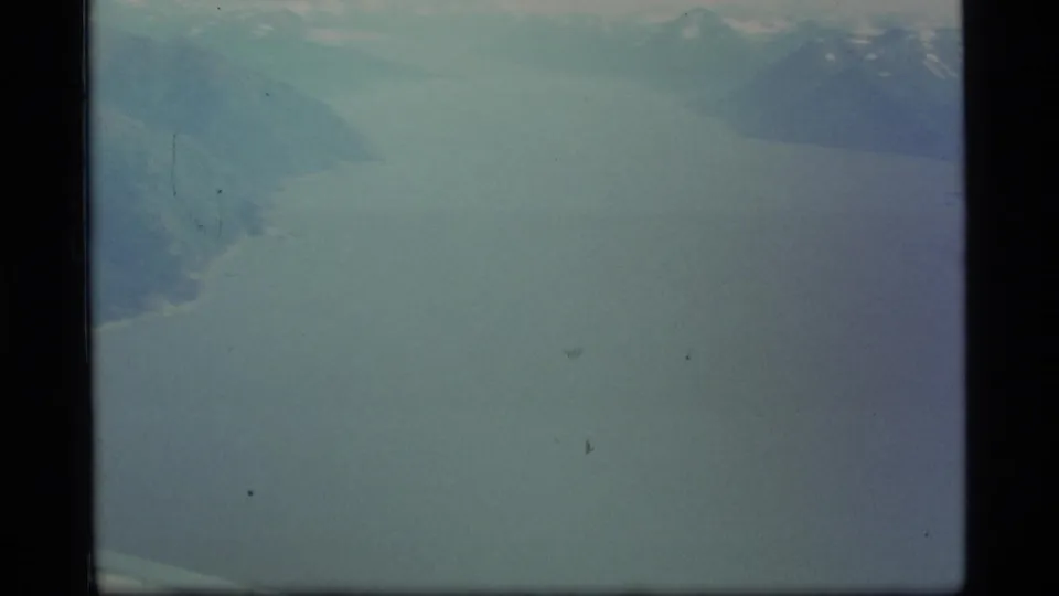 1977 8mm Alaska aerial frame with pale blue water or haze and darker mountainous or shoreline forms near the upper edge.