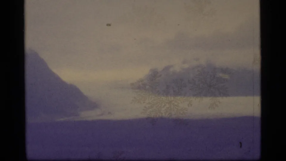 1977 Alaska home-movie frame of distant snowy mountains and hazy water under cloud cover, tinted purple by the film scan.