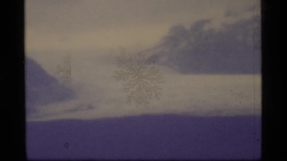 1977 8mm Alaska frame showing a misty mountain shoreline with surface snowflake marks and muted purple-blue tones.
