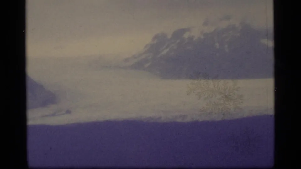 1977 Alaska 8mm frame with a snowy mountain ridge, pale water or ice, cloudy sky, and visible film grain.