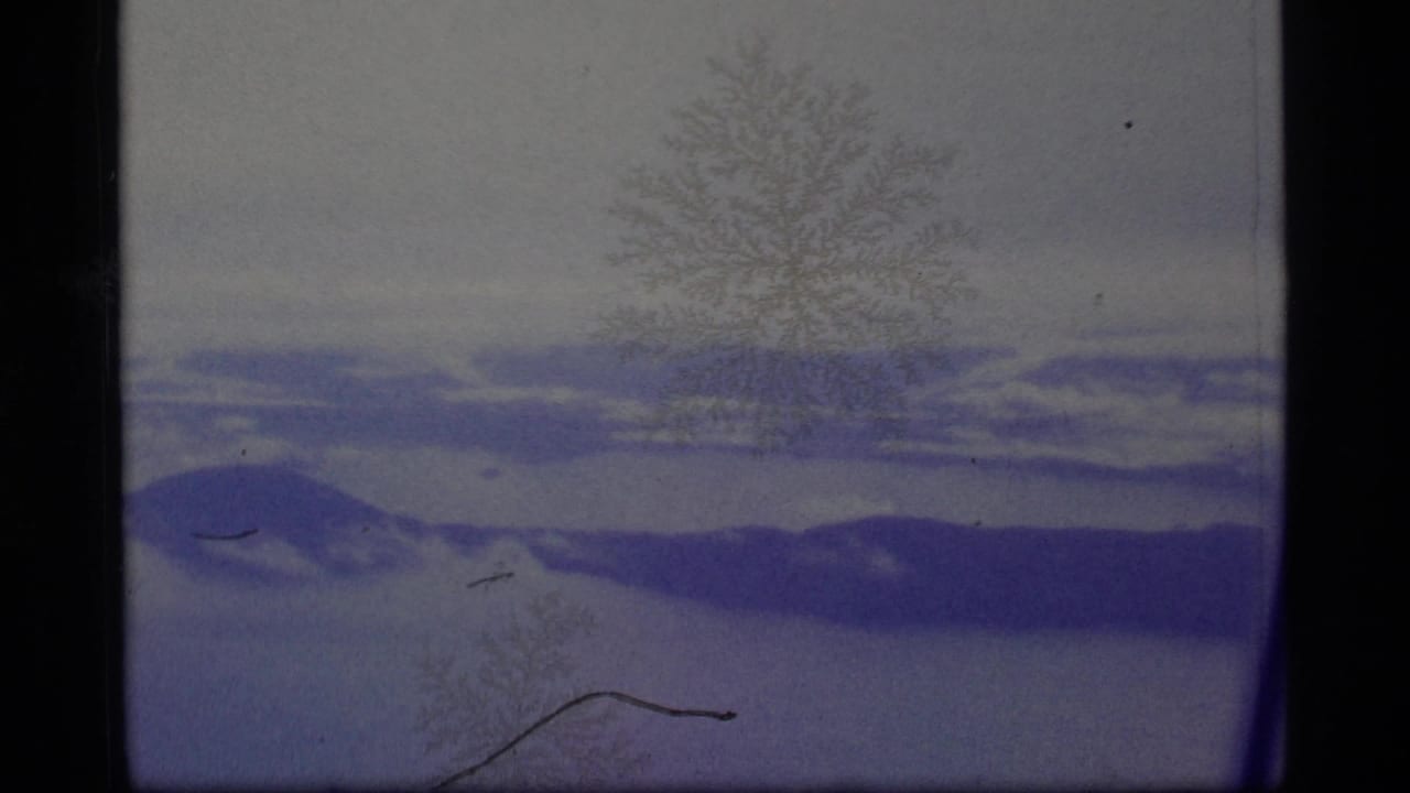 1977 8mm Alaska winter frame showing snow-covered terrain, distant dark ridges, pale sky, and frost-like marks across the film image.