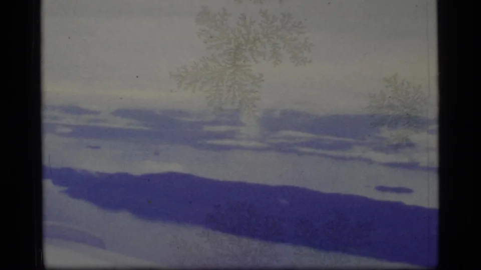 1977 8mm Alaska winter frame showing snow-covered terrain, blue-purple ridge forms, pale sky, and soft crystal-like marks on the image.