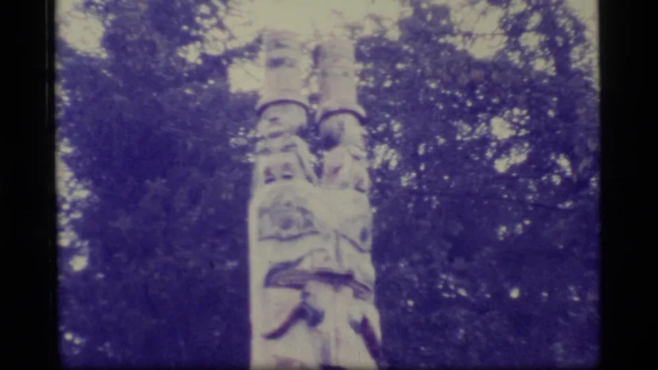A tall carved totem pole appears among dense trees in 1977 Alaska home-movie footage with a purple color cast.