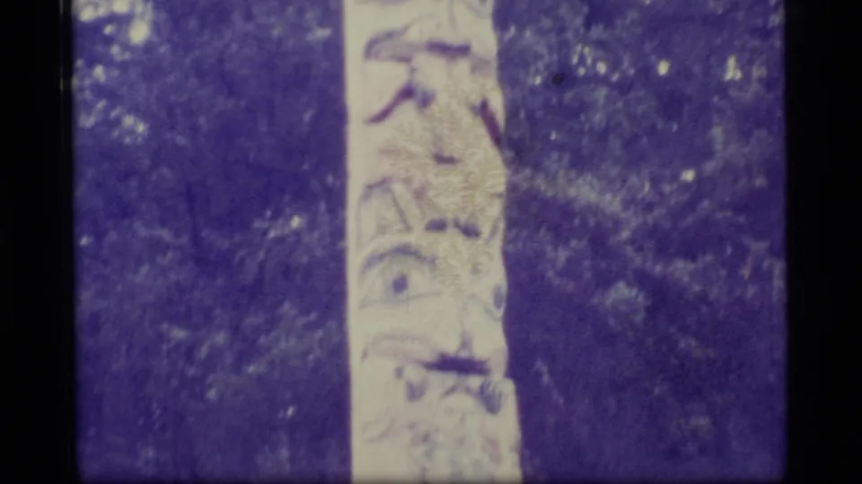 A closer 1977 Alaska Super 8 view shows carved details on the lower portion of a totem pole surrounded by trees.