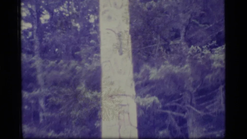 1977 Alaska 8mm frame with the upper portion of a carved totem pole against dense evergreen foliage and aged color shift.