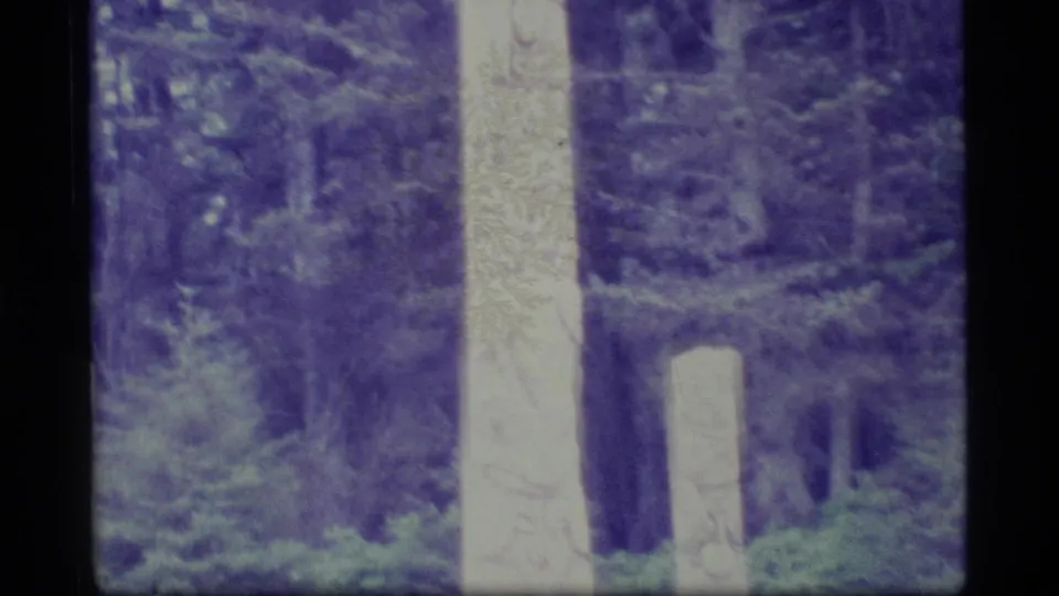 1977 Alaska 8mm frame showing the carved pole in a forest clearing area with evergreens and a pale stump or post nearby.