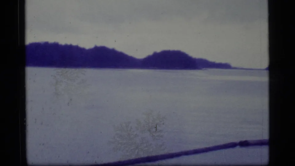 1977 Alaska 8mm frame showing calm coastal water, distant wooded hills, and a dark shoreline edge with faded color.